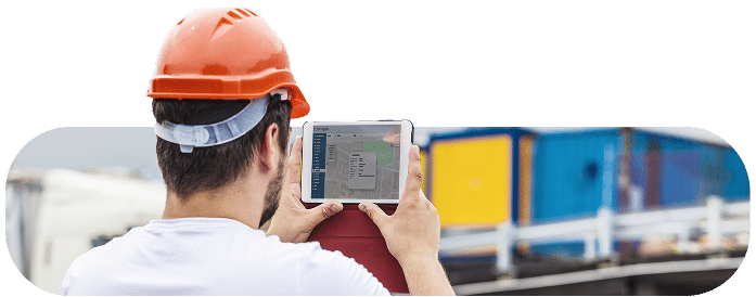 Un ouvrier portant un casque de sécurité orange utilise une tablette pour consulter une application de gestion de chantier sur site, devant des équipements.