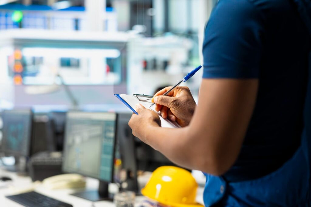 Technicien en blouse bleue effectuant une maintenance préventive, prenant des notes sur un clipboard devant des ordinateurs et un casque de sécurité jaune.