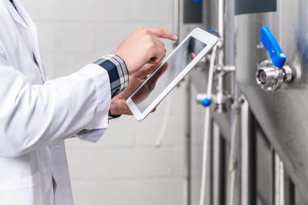 Ingénieur seul dans une usine de traitement (station ou brasserie) entourée de vannes et de tuyaux, consultant les données sur une tablette.